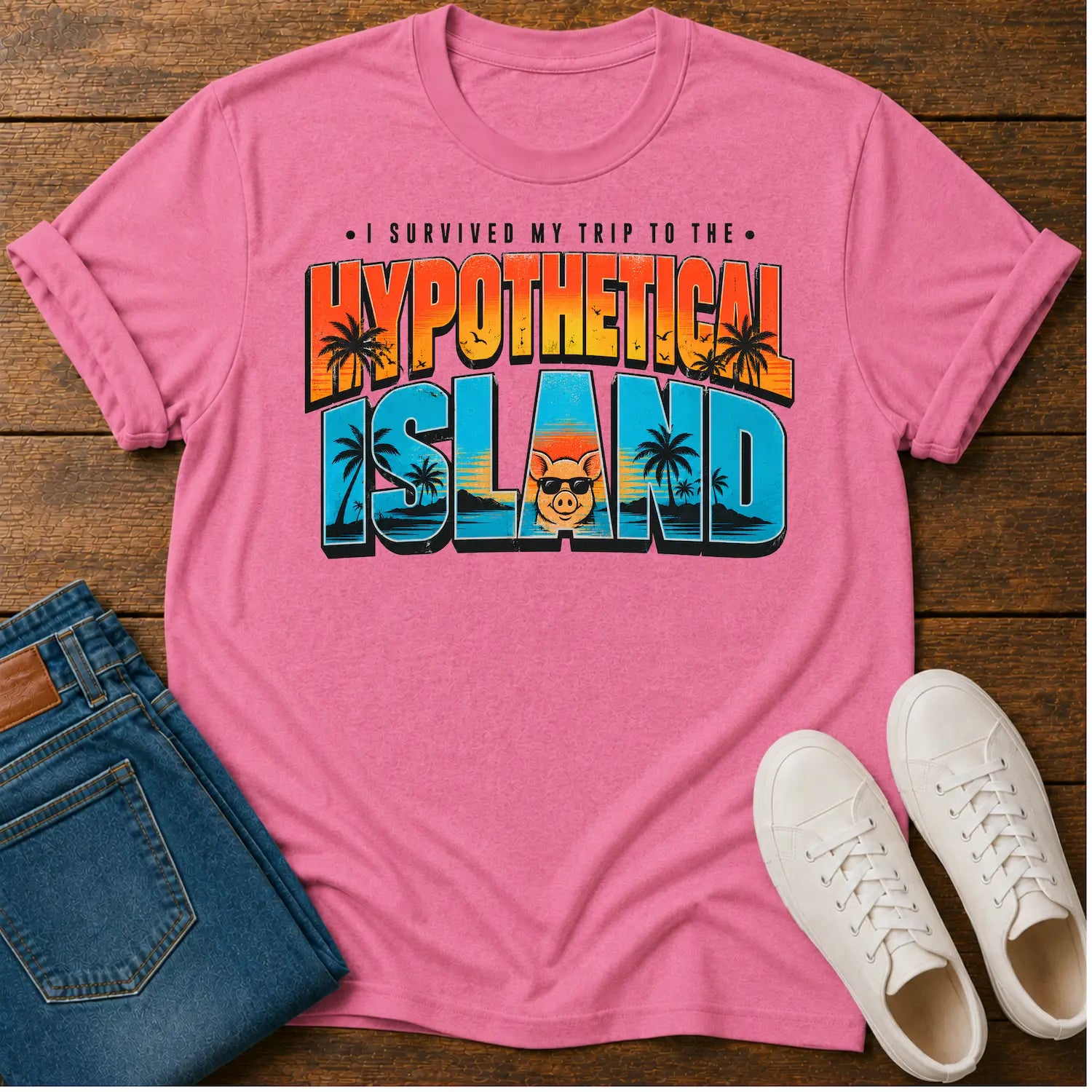 A pink short-sleeve graphic T-shirt laid flat on a wooden surface, styled with blue jeans and white sneakers. The shirt features a colorful retro-style text graphic reading 'I SURVIVED MY TRIP TO THE HYPOTHETICAL ISLAND' with palm trees and a cartoon face.