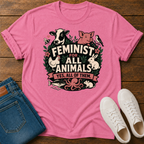 A pink graphic t-shirt laid flat on a wooden surface, styled with folded blue jeans and white sneakers. The t-shirt features a colorful graphic of various animals (cow, pig, chicken, rabbit, octopus, fish) surrounding the text 'FEMINIST FOR ALL ANIMALS YES, ALL OF THEM.
