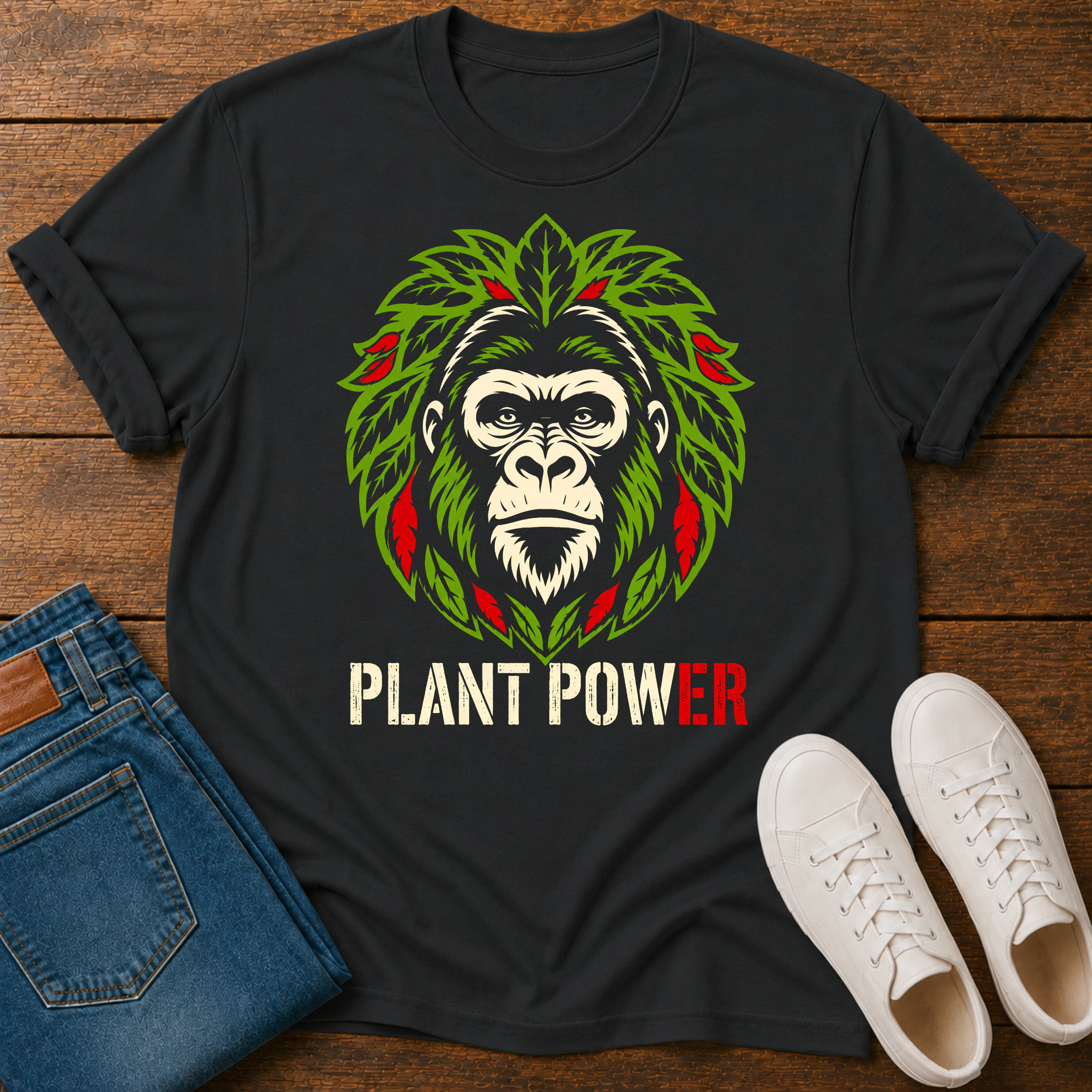 A black graphic t-shirt laid flat on a wooden surface, styled with blue jeans and white sneakers. The t-shirt features the text 'plant power' and a gorilla head graphic