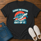 A black graphic t-shirt laid flat on a wooden surface, styled with folded blue jeans and white sneakers. The t-shirt features a retro-style graphic of a sea turtle swimming in front of a red sun over blue waves, with the text 'LEAVE THE OCEAN OUT OF IT'.