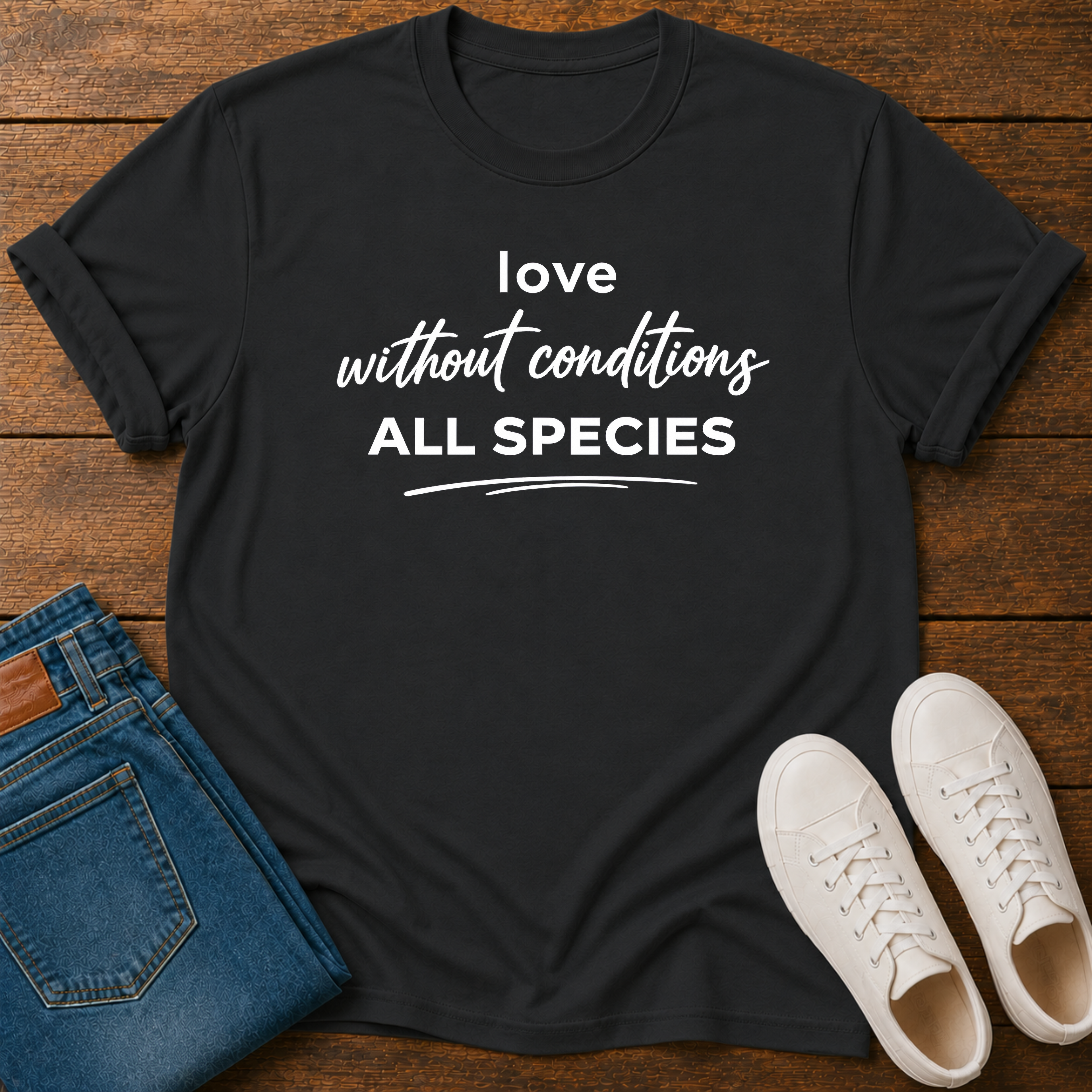 A black short-sleeve graphic T-shirt laid flat on a wooden surface, styled with folded blue jeans and a pair of white sneakers. The shirt has white text reading 'love without conditions ALL SPECIES.