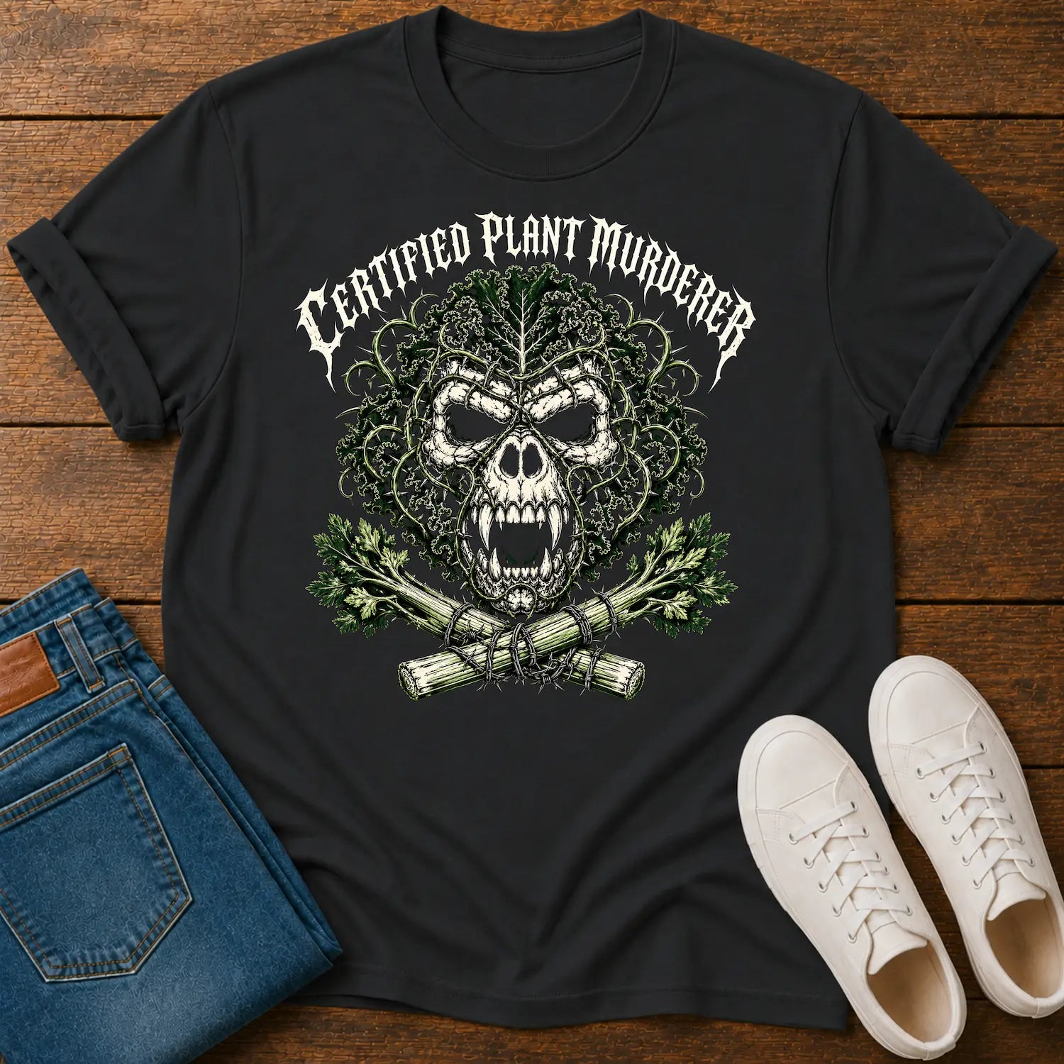 A black graphic t-shirt featuring a skull made of leaves and branches with the text 'CERTIFIED PLANT MURDERER' above it, styled in a flat lay on a wooden surface with blue jeans and white sneakers.