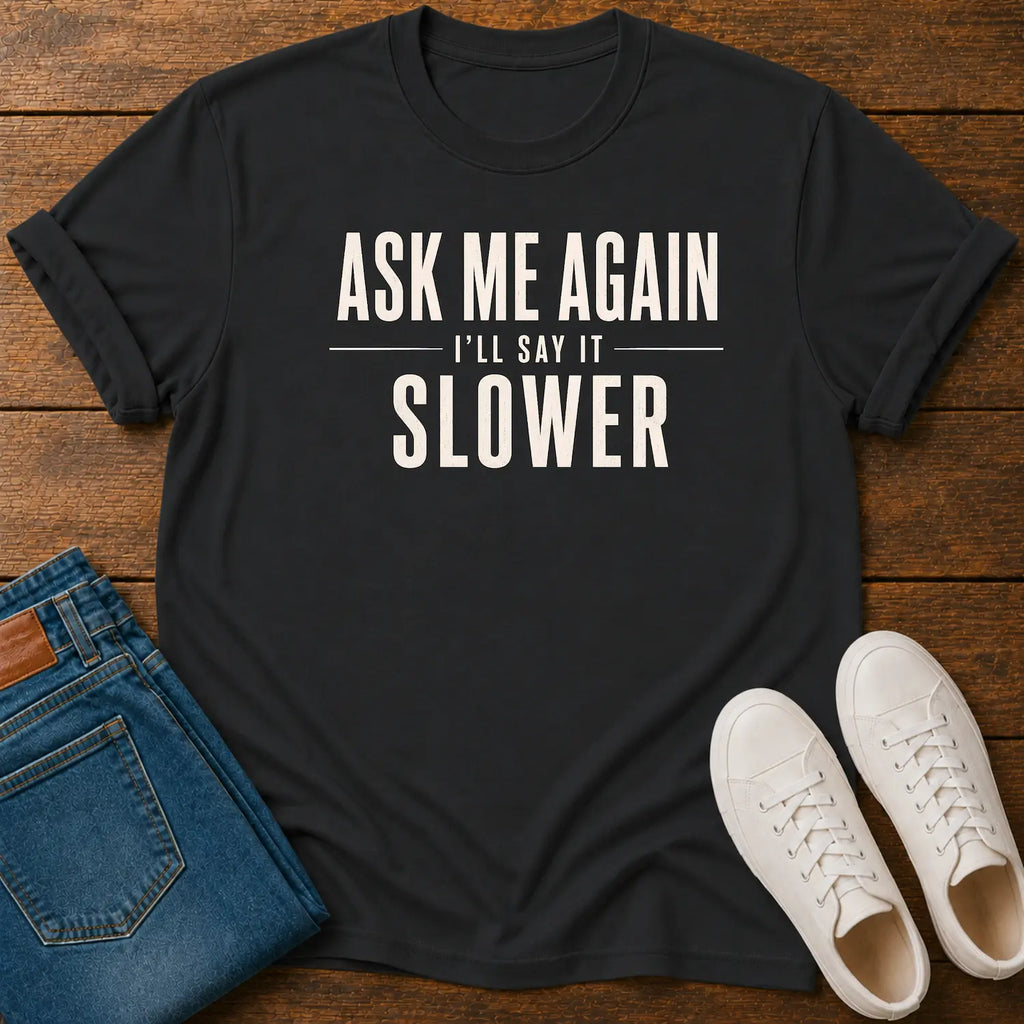 A black graphic t-shirt with the text 'ASK ME AGAIN I'LL SAY IT SLOWER' in white, styled in a flat lay on a wooden background with blue jeans and white sneakers.