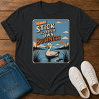 A black graphic t-shirt laid flat on a wooden surface, styled with blue jeans and white sneakers. The graphic features a cartoon duck in a pond with the text 'PLEASE STICK TO YOUR OWN BUSINESS'.