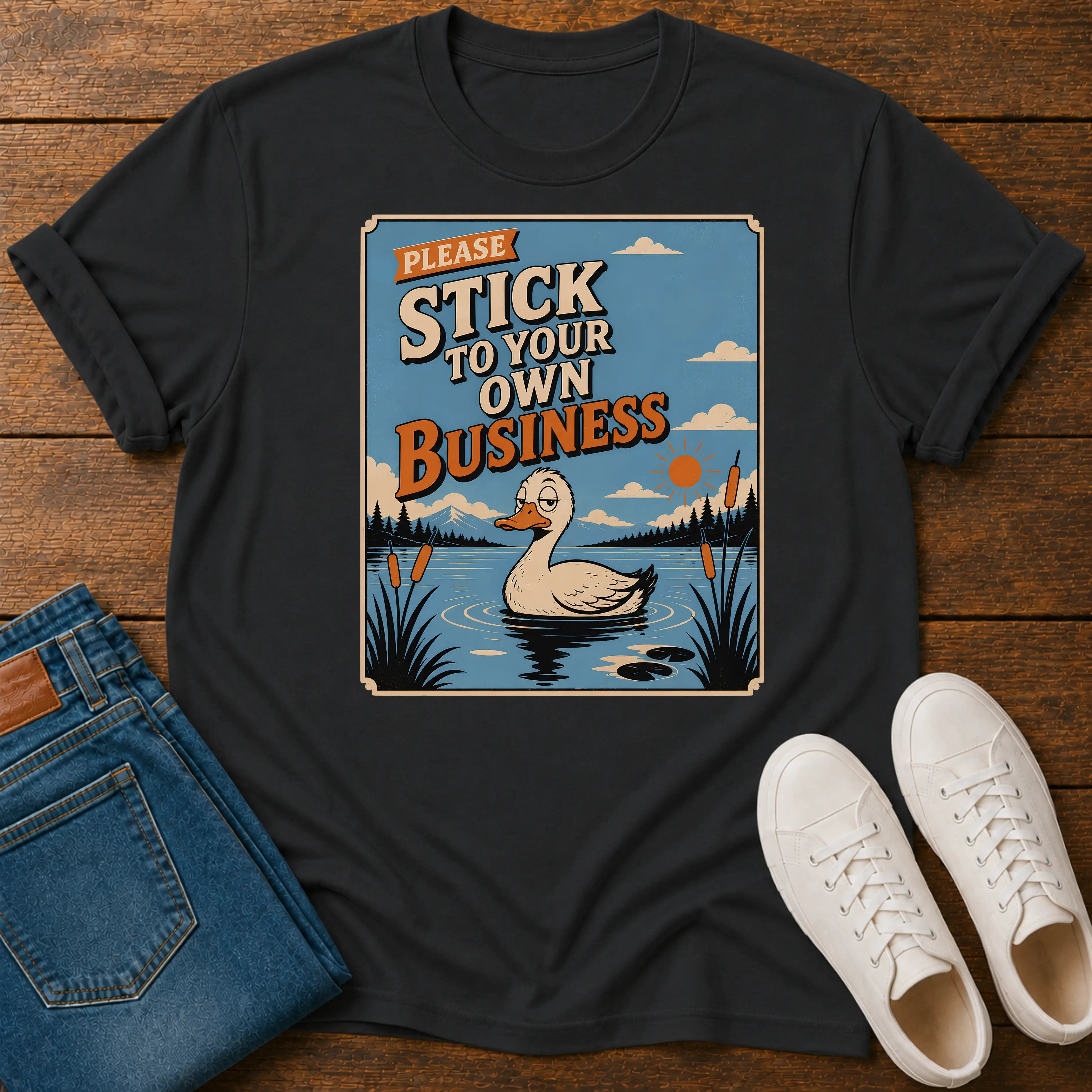 A black graphic t-shirt laid flat on a wooden surface, styled with blue jeans and white sneakers. The graphic features a cartoon duck in a pond with the text 'PLEASE STICK TO YOUR OWN BUSINESS'.
