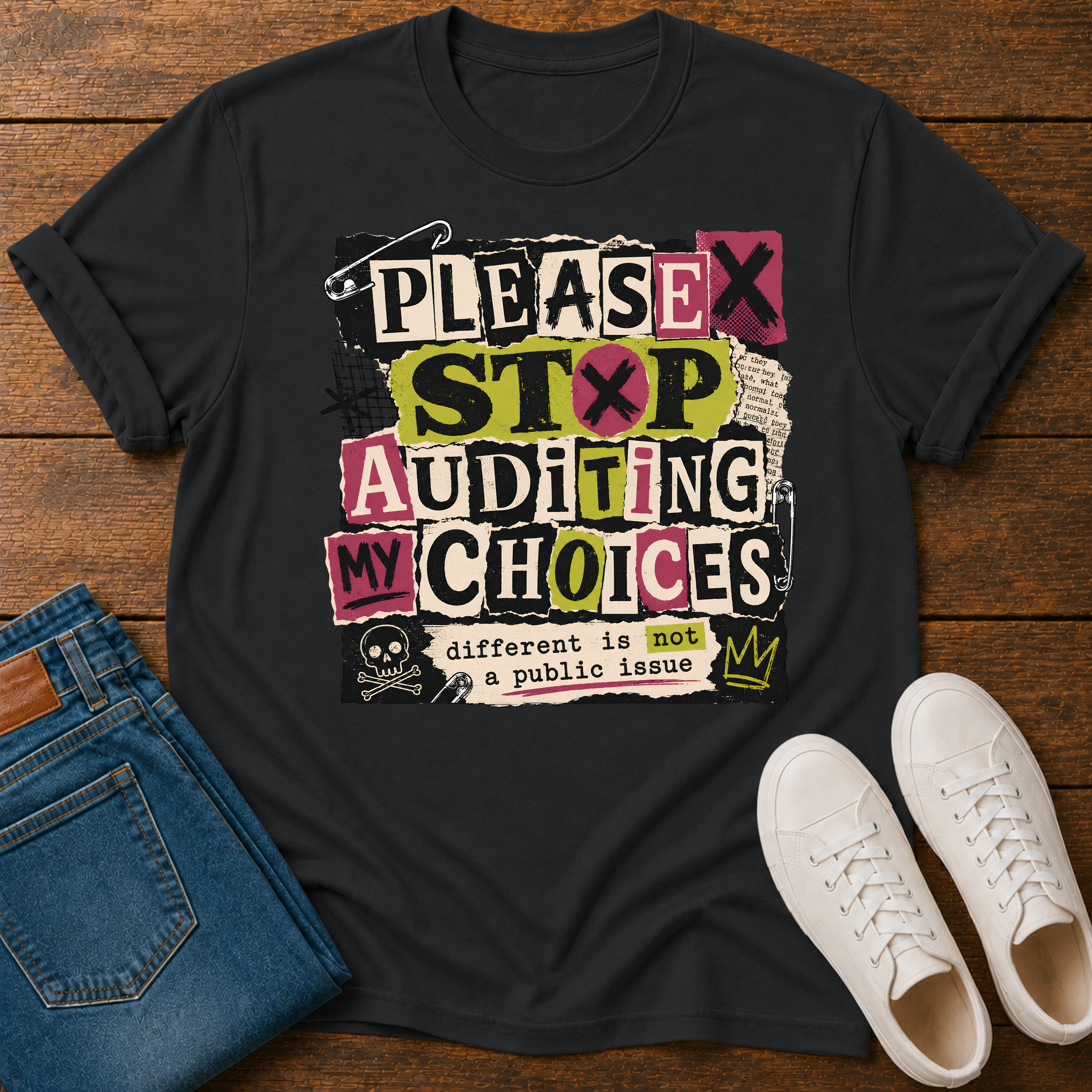 A black graphic t-shirt laid flat on a wooden surface, styled with folded blue jeans and white sneakers. The t-shirt features a bold, multi-colored graphic with the text 'PLEASE STOP AUDITING MY CHOICES' and 'different is not a public issue' with a small crown and skull graphic.