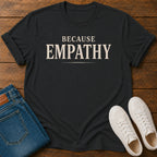 A black-shirt with the word 'EMPATHY' printed in large cream-colored letters, styled in a flat lay on a wooden background alongside folded blue jeans and a pair of white sneakers.