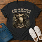 A black graphic t-shirt laid flat on a wooden surface, styled with folded blue jeans and white sneakers. The t-shirt features a sepia-toned graphic of an elephant walking through a jungle path, with the text 'JUST BECAUSE SOMETHING EXISTS DOES NOT MEAN IT BELONGS TO YOU' printed above the graphic.