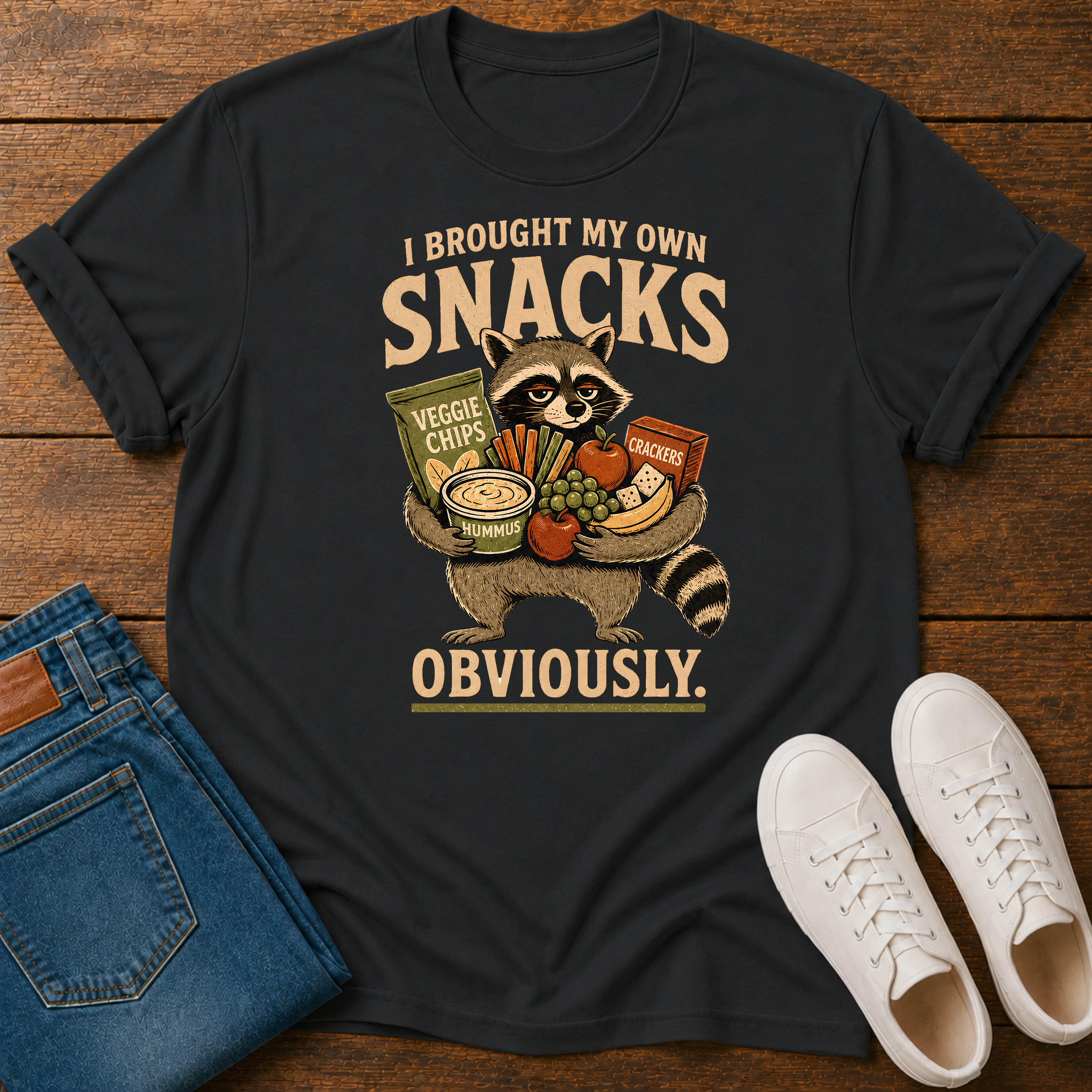 A black graphic t-shirt laid flat on a wooden surface, styled with a pair of blue jeans and white sneakers. The t-shirt features a cartoon raccoon holding various snacks with the text 'I BROUGHT MY OWN SNACKS OBVIOUSLY.