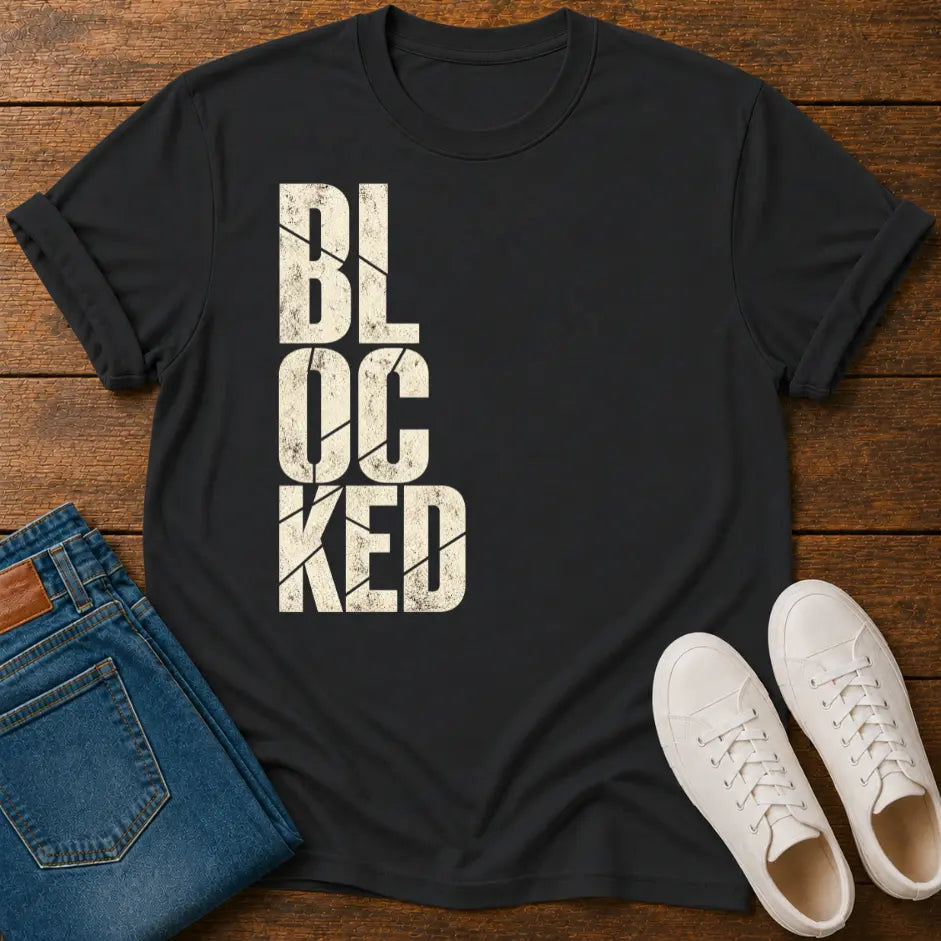 A black short-sleeve t-shirt with the word 'BLOCKED' printed vertically in distressed off-white block letters, styled in a flat lay on a wooden surface alongside blue jeans and white sneakers.