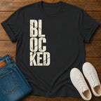 A black short-sleeve t-shirt with the word 'BLOCKED' printed vertically in distressed off-white block letters, styled in a flat lay on a wooden surface alongside blue jeans and white sneakers.