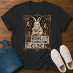 A black graphic t-shirt laid flat on a wooden surface, styled with folded blue jeans and white sneakers. The t-shirt features a vintage-style illustration of a rabbit holding a sign that says 'NO' surrounded by birds, mushrooms, and the text 'ASK someone ELSE'.