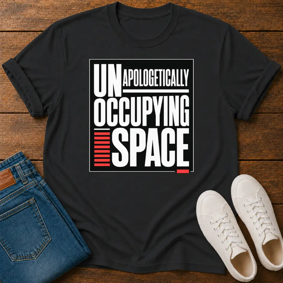 A black short-sleeve graphic T-shirt laid flat on a wooden surface, styled with folded blue jeans and a pair of white sneakers. The shirt has a large front print reading “UNAPOLOGETHICALLY OCCUPING SPACE” in white, with red graphic accents.