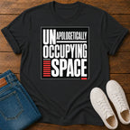 A black short-sleeve graphic T-shirt laid flat on a wooden surface, styled with folded blue jeans and a pair of white sneakers. The shirt has a large front print reading “UNAPOLOGETHICALLY OCCUPING SPACE” in white, with red graphic accents.