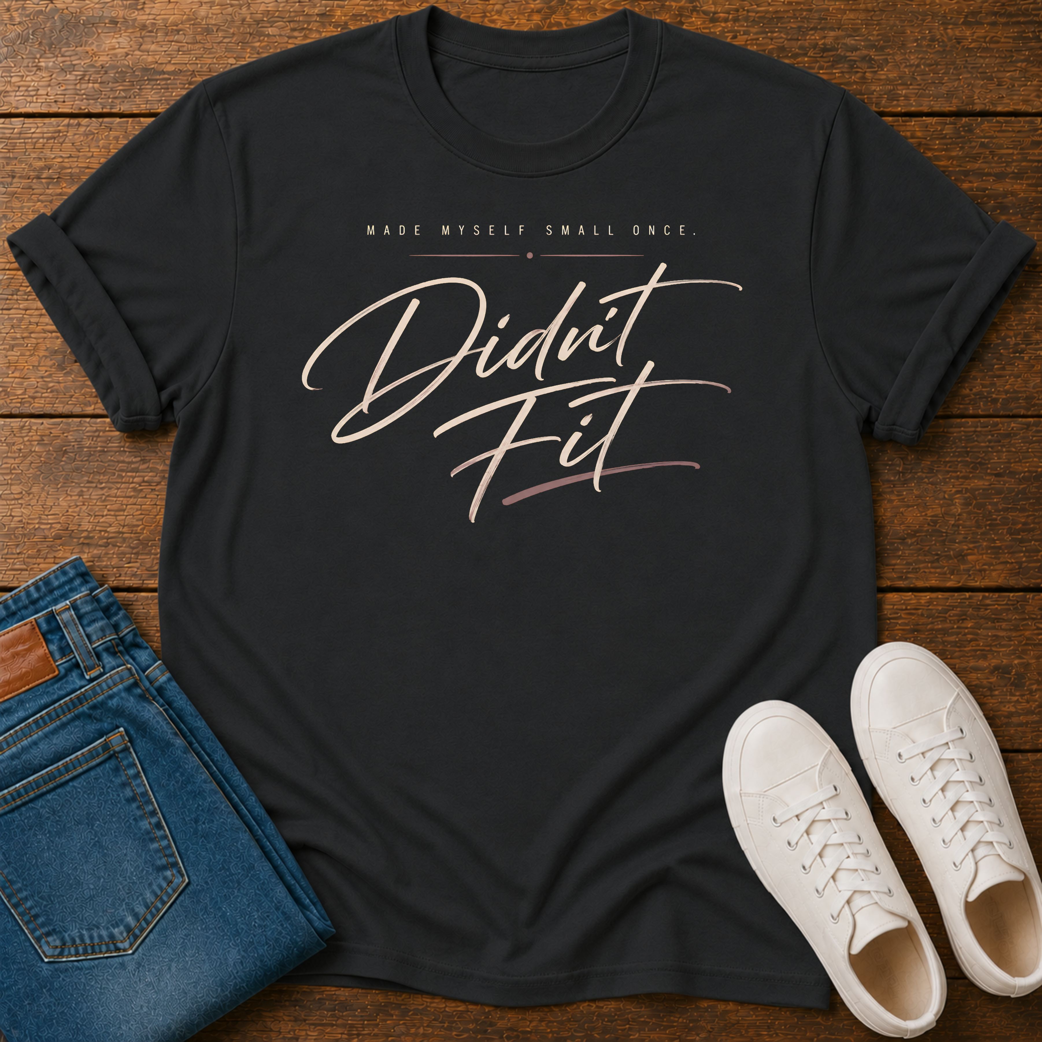A black graphic t-shirt laid flat on a wooden surface, styled with folded blue jeans and a pair of white sneakers. The t-shirt features the text 'MADE MYSELF SMALL ONCE.