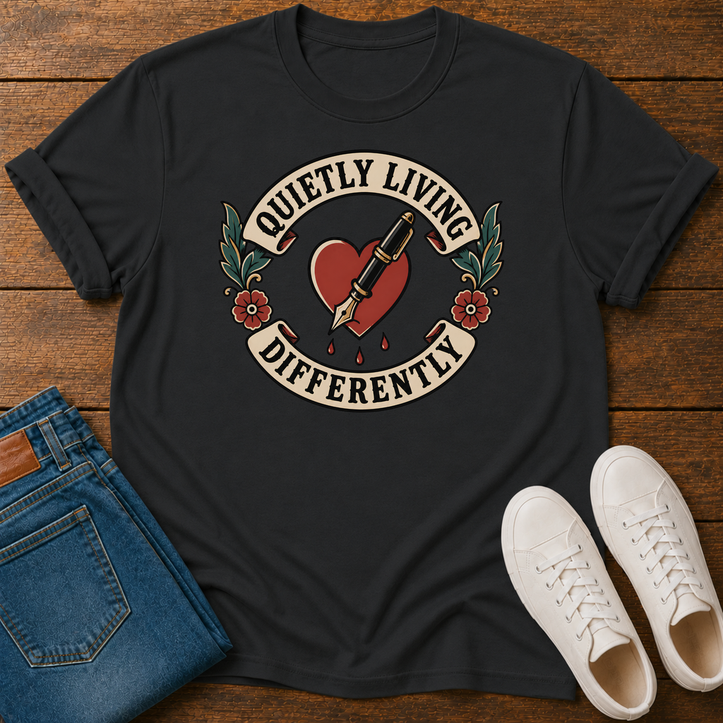 A black graphic t-shirt laid flat on a wooden surface, styled with folded blue jeans and white sneakers. The t-shirt features a retro tattoo-style graphic of a red heart pierced by a gold pen, with the text 'QUIETLY LIVING DIFFERENTLY' arched above and below.