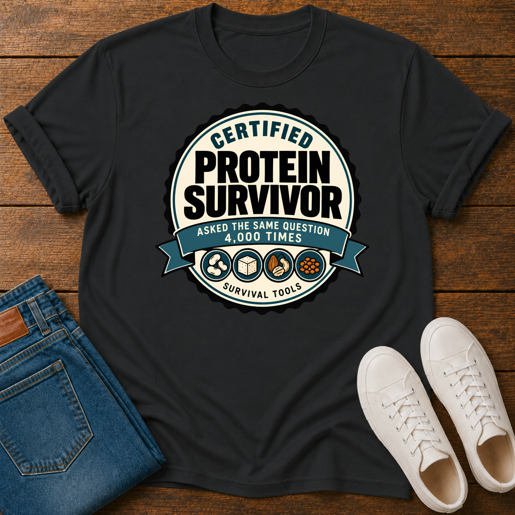 A black graphic t-shirt laid flat on a wooden surface, styled with folded blue jeans and white sneakers. The t-shirt features a circular graphic that reads 'CERTIFIED PROTEIN SURVIVOR' with 'ASKED THE SAME QUESTION 4,000 TIMES' and 'SURVIVAL TOOLS' on a blue ribbon banner.