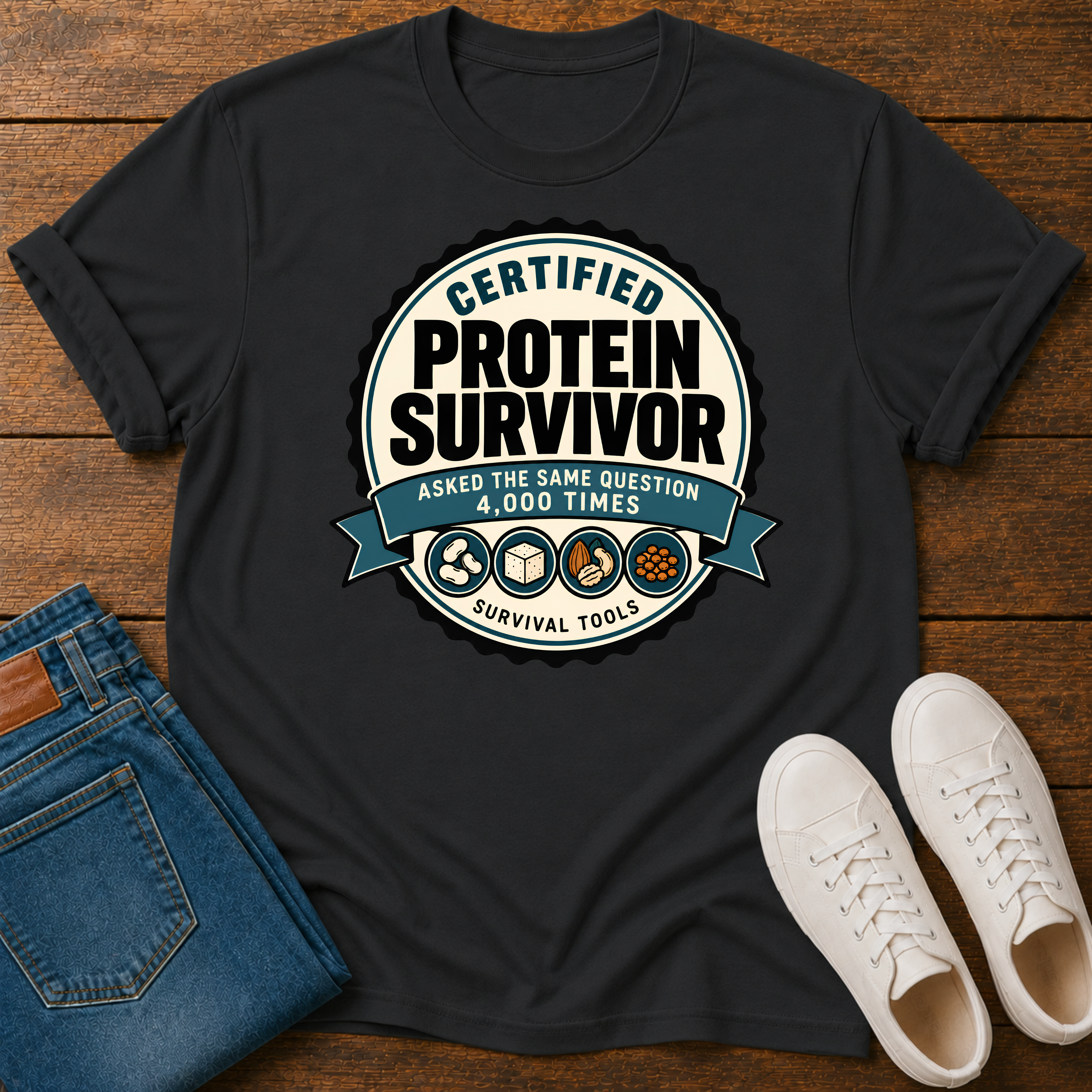 A black graphic t-shirt laid flat on a wooden surface, styled with folded blue jeans and white sneakers. The t-shirt features a circular graphic that reads 'CERTIFIED PROTEIN SURVIVOR' with 'ASKED THE SAME QUESTION 4,000 TIMES' and 'SURVIVAL TOOLS' on a blue ribbon banner.