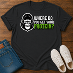 A black short-sleeve graphic T-shirt laid flat on a wooden surface, styled with folded blue jeans and white sneakers. The shirt features a large gorilla skull design with leaf details, crossed plant stems, and the text ''where do you get your protein from?"