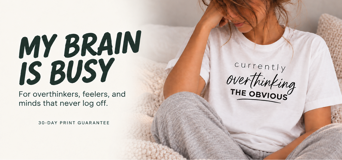 A woman sitting on a couch wearing a white graphic t-shirt that reads 'currently overthinking THE OBVIOUS' in black script and serif text. The image has a large text overlay on the left reading 'MY BRAIN IS BUSY' with supporting text about overthinkers and a '30-DAY PRINT GUARANTEE'.