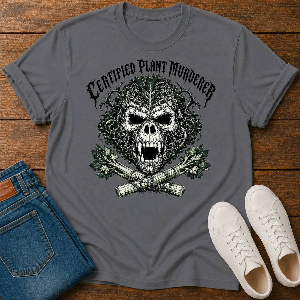 A dark gray graphic t-shirt featuring a skull made of leaves and branches with crossed plants underneath and the text 'CERTIFIED PLANT MURDERER' above. The t-shirt is styled in a flat-lay on a wooden surface alongside blue jeans and white sneakers.
