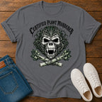 A dark gray graphic t-shirt featuring a skull made of leaves and branches with crossed plants underneath and the text 'CERTIFIED PLANT MURDERER' above. The t-shirt is styled in a flat-lay on a wooden surface alongside blue jeans and white sneakers.