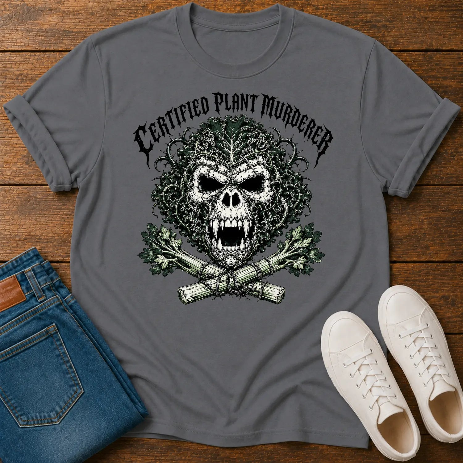 A dark gray graphic t-shirt featuring a skull made of leaves and branches with crossed plants underneath and the text 'CERTIFIED PLANT MURDERER' above. The t-shirt is styled in a flat-lay on a wooden surface alongside blue jeans and white sneakers.