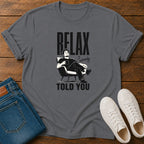 A dark gray graphic t-shirt laid flat on a wooden surface, styled with folded blue jeans and white sneakers. The t-shirt features a black graphic of a man relaxing in a chair with the text 'RELAX I'm ready TOLD YOU'.