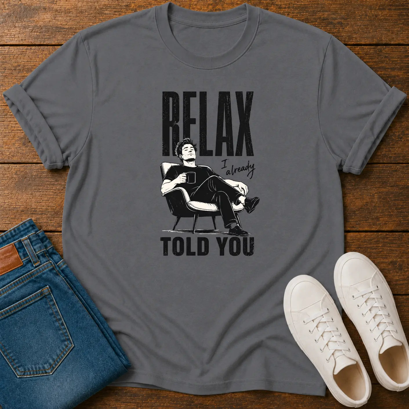 A dark gray graphic t-shirt laid flat on a wooden surface, styled with folded blue jeans and white sneakers. The t-shirt features a black graphic of a man relaxing in a chair with the text 'RELAX I'm ready TOLD YOU'.