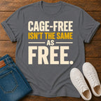 A dark gray graphic t-shirt laid flat on a wooden surface, styled with folded blue jeans and white sneakers. The t-shirt features the text 'CAGE-FREE ISN'T THE SAME AS FREE.