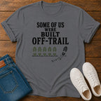 A gray graphic t-shirt laid flat on a wooden surface, styled with folded blue jeans and a pair of white sneakers. The t-shirt features the black text 'SOME OF US WERE BUILT OFF-TRAIL' above green boot prints and a black footprint trail graphic.