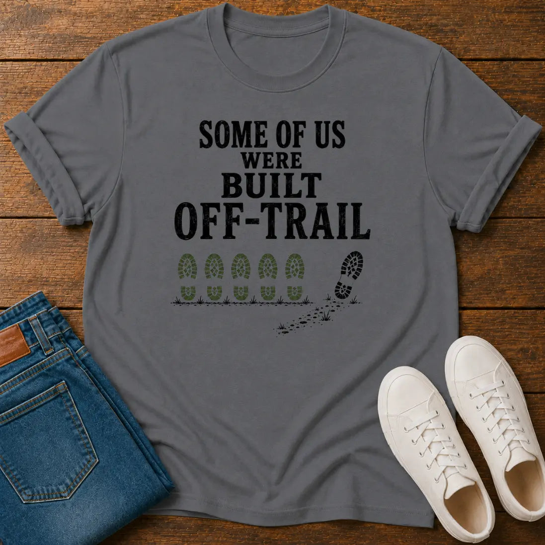 A gray graphic t-shirt laid flat on a wooden surface, styled with folded blue jeans and a pair of white sneakers. The t-shirt features the black text 'SOME OF US WERE BUILT OFF-TRAIL' above green boot prints and a black footprint trail graphic.