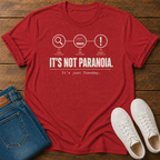 Cherry red short-sleeve graphic T-shirt laid flat on a wooden surface, styled with folded blue jeans and white sneakers.  text 'it's not paranoia it's a tuesday'