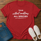 A red short-sleeve graphic T-shirt laid flat on a wooden surface, styled with folded blue jeans and a pair of white sneakers. The shirt has white text reading 'love without conditions ALL SPECIES.