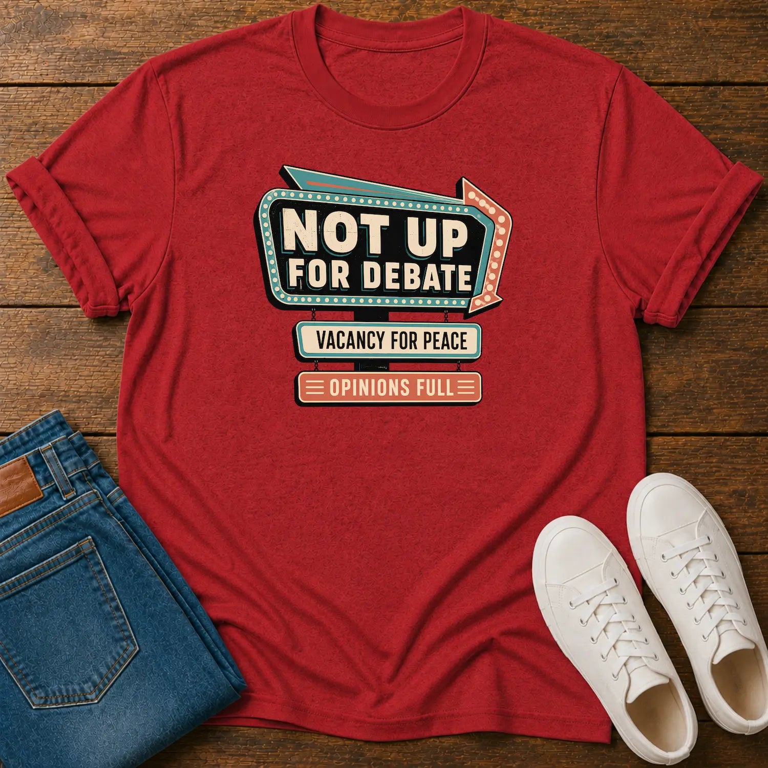A red graphic t-shirt laid flat on a wooden surface, styled with folded blue jeans and a pair of white sneakers. The t-shirt features a retro marquee-style graphic that reads 'NOT UP FOR DEBATE' with 'VACANCY FOR PEACE' and 'OPINIONS FULL' underneath.