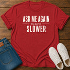 A red graphic t-shirt with the text 'ASK ME AGAIN I'LL SAY IT SLOWER' in white, styled in a flat lay on a wooden background with blue jeans and white sneakers.