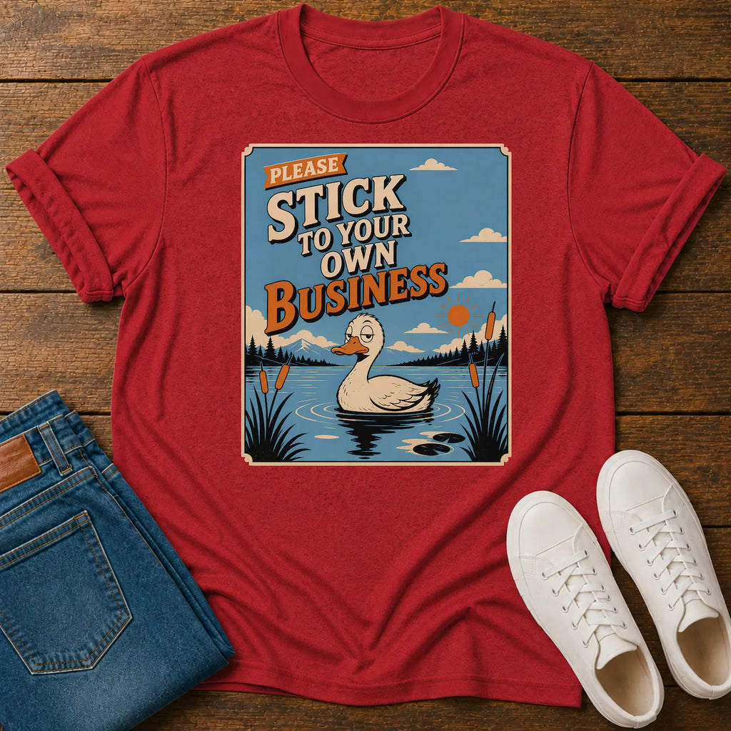 A red graphic t-shirt laid flat on a wooden surface, styled with folded blue jeans and white sneakers. The t-shirt features a square graphic with a duck in a lake and the text 'PLEASE STICK TO YOUR OWN BUSINESS'.