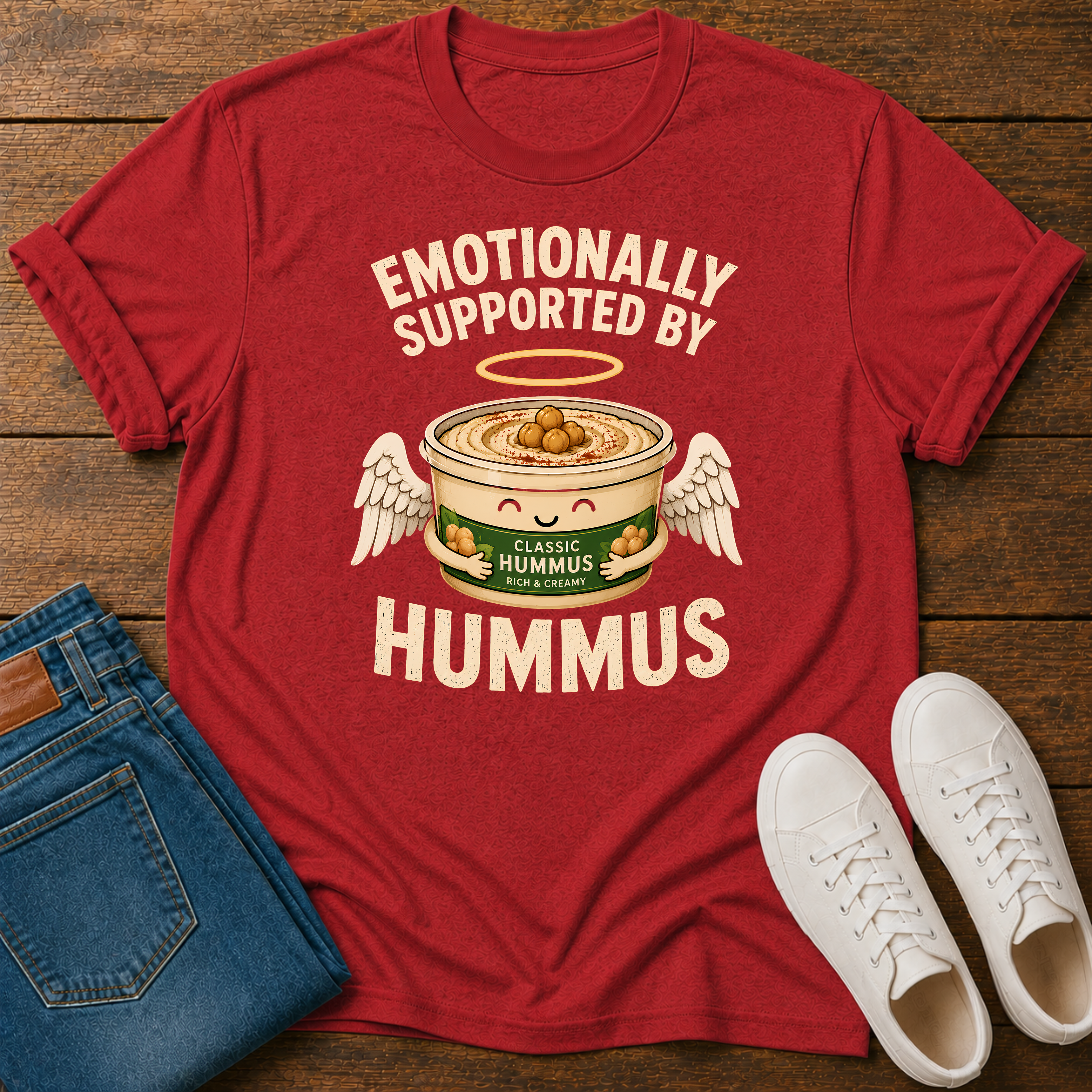 A red graphic t-shirt laid flat on a wooden surface, styled with folded blue jeans and a pair of white sneakers. The t-shirt features a humorous graphic of a can of hummus with angel wings and a halo, with the text 'EMOTIONALLY SUPPORTED BY HUMMUS'.