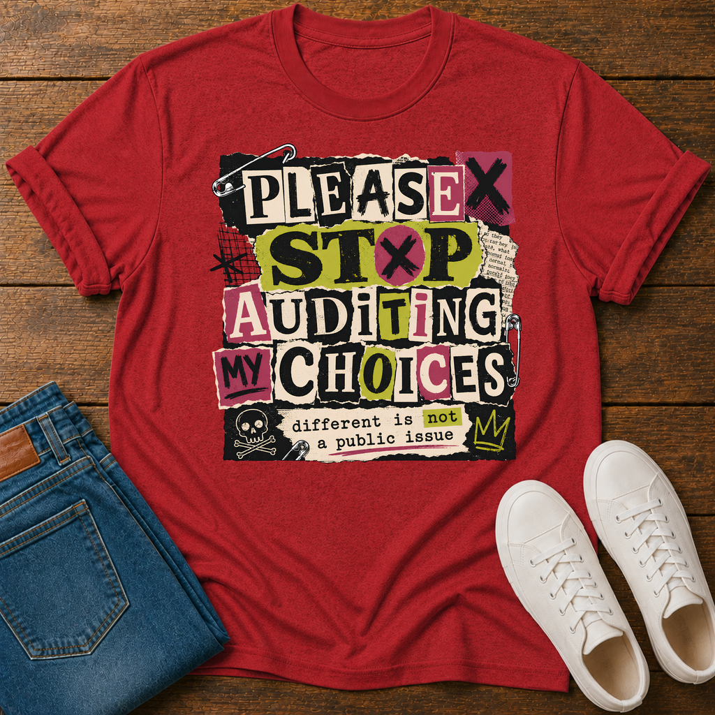A red graphic t-shirt laid flat on a wooden surface, styled with folded blue jeans and white sneakers. The t-shirt features a bold, multi-colored text graphic reading 'PLEASE STOP AUDITING MY CHOICES' with a skull and crown detail.
