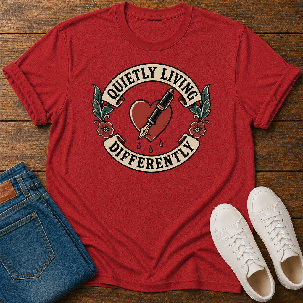 A red graphic t-shirt laid flat on a wooden surface, styled with folded blue jeans and a pair of white sneakers. The t-shirt features a vintage-style tattoo design with a heart, fountain pen, and the text 'QUIETLY LIVING DIFFERENTLY'.