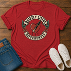 A red graphic t-shirt laid flat on a wooden surface, styled with folded blue jeans and a pair of white sneakers. The t-shirt features a vintage-style tattoo design with a heart, fountain pen, and the text 'QUIETLY LIVING DIFFERENTLY'.