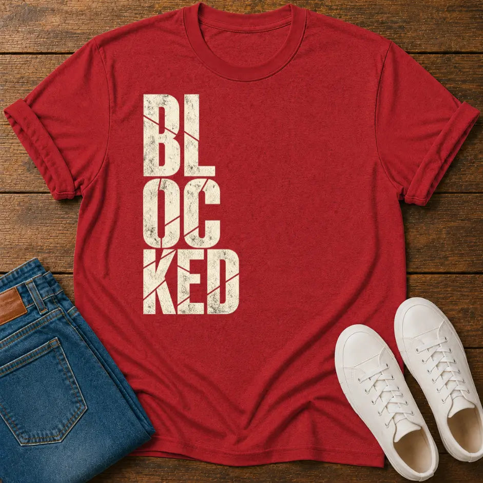 A red short-sleeve t-shirt with the word 'BLOCKED' printed vertically in distressed white block letters, styled in a flat lay on a wooden surface with blue jeans and white sneakers.