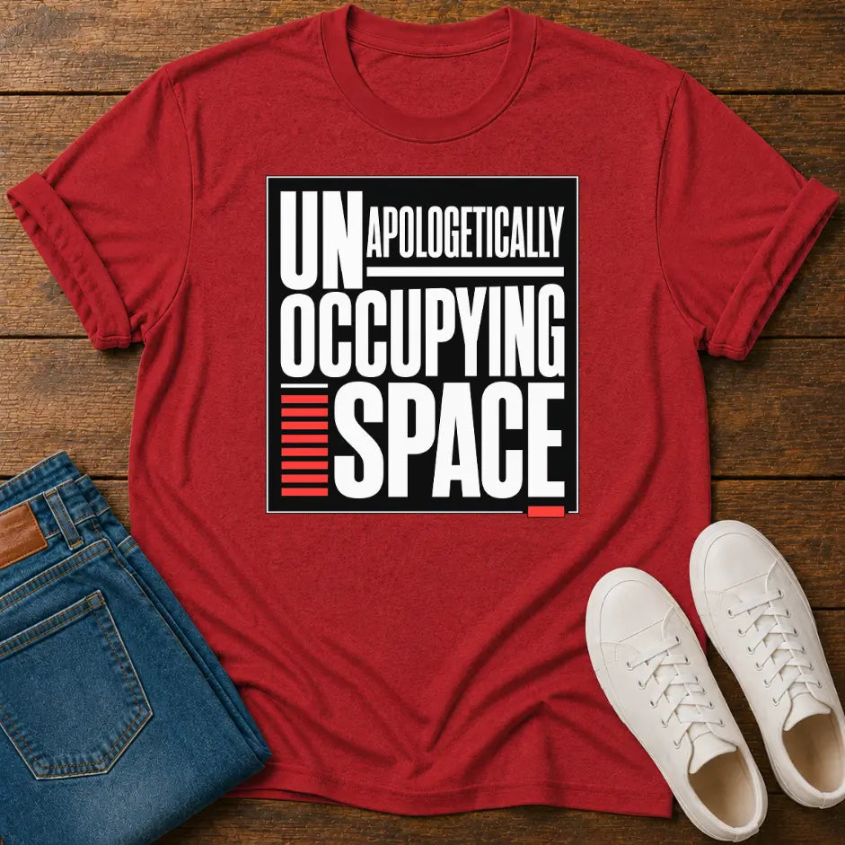 A red short-sleeve graphic T-shirt laid flat on a wooden surface, styled with folded blue jeans and white sneakers. The shirt has a large black-and-white text graphic reading 'UNAPOLOGETICALLY OCCUPYING SPACE' with red accent bars.