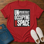 A red short-sleeve graphic T-shirt laid flat on a wooden surface, styled with folded blue jeans and white sneakers. The shirt has a large black-and-white text graphic reading 'UNAPOLOGETICALLY OCCUPYING SPACE' with red accent bars.