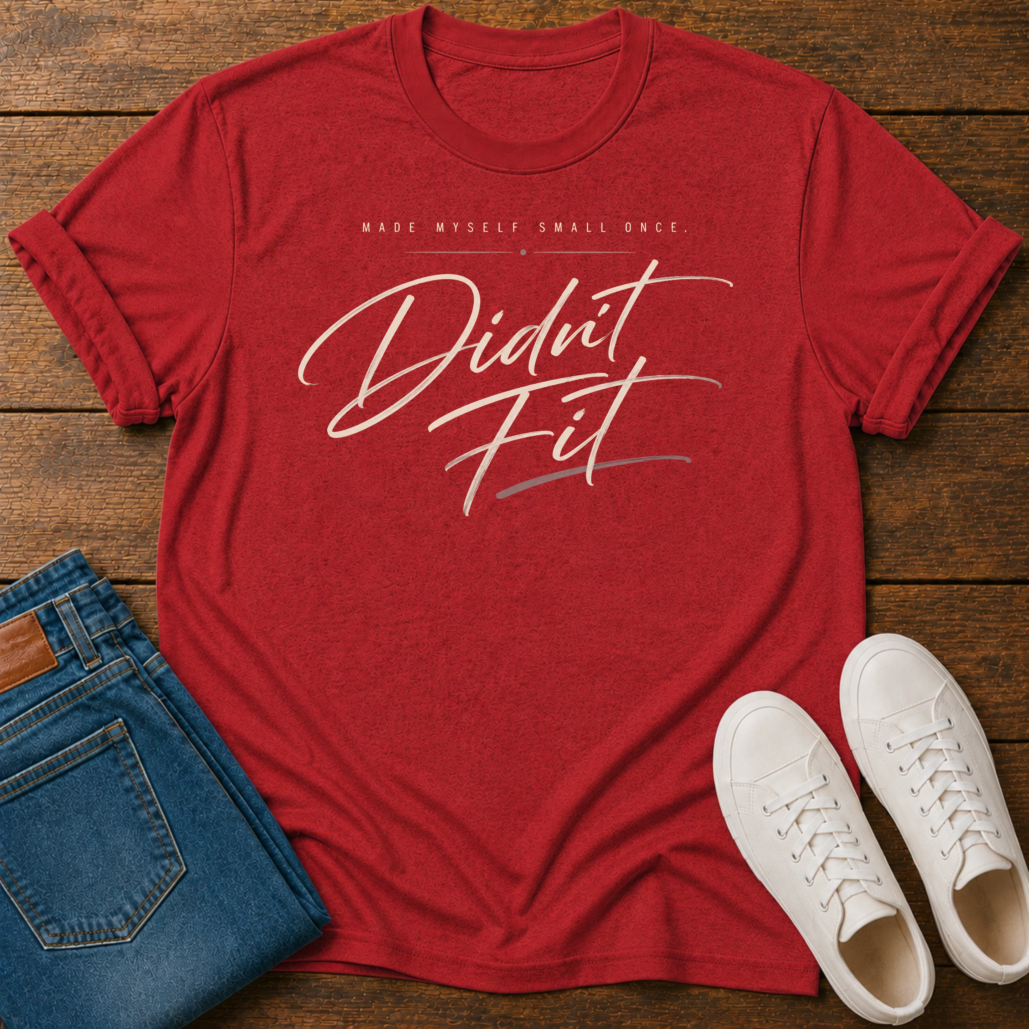 A red graphic t-shirt laid flat on a wooden surface, styled with folded blue jeans and white sneakers. The t-shirt features the text 'MADE MYSELF SMALL ONCE. DIDN'T FIT'