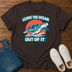 A dark brown graphic t-shirt laid flat on a wooden surface, styled with folded blue jeans and white sneakers. The t-shirt features a retro-style graphic of a sea turtle swimming in front of a red and orange sun, with the text 'LEAVE THE OCEAN OUT OF IT' in white lettering on blue banners.