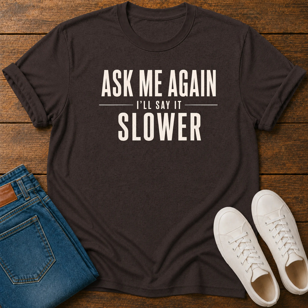 A dark charcoal gray graphic t-shirt with the text 'ASK ME AGAIN I'LL SAY IT SLOWER' in cream lettering, styled in a flat lay on a wooden background alongside folded blue jeans and white sneakers.