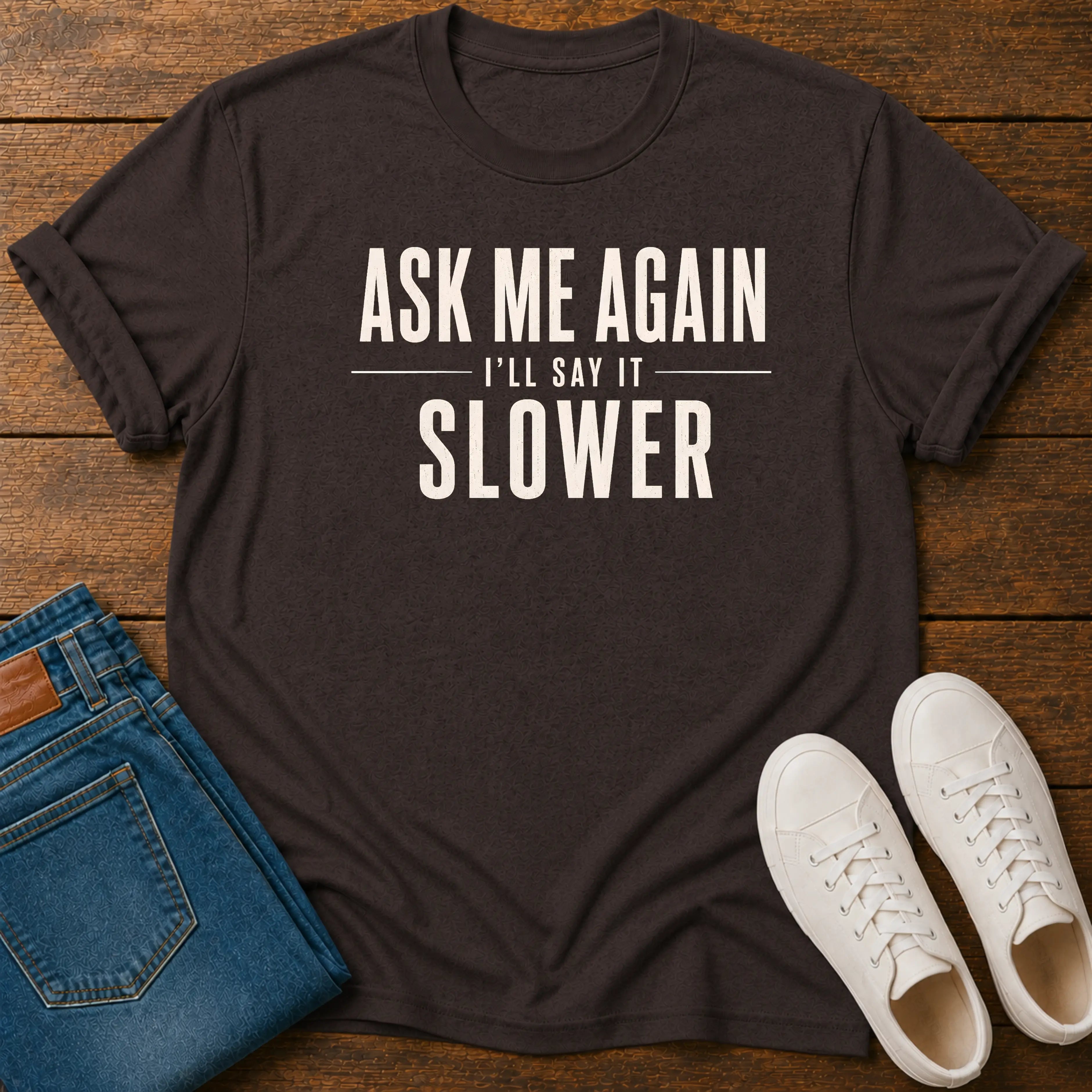 A dark charcoal gray graphic t-shirt with the text 'ASK ME AGAIN I'LL SAY IT SLOWER' in cream lettering, styled in a flat lay on a wooden background alongside folded blue jeans and white sneakers.