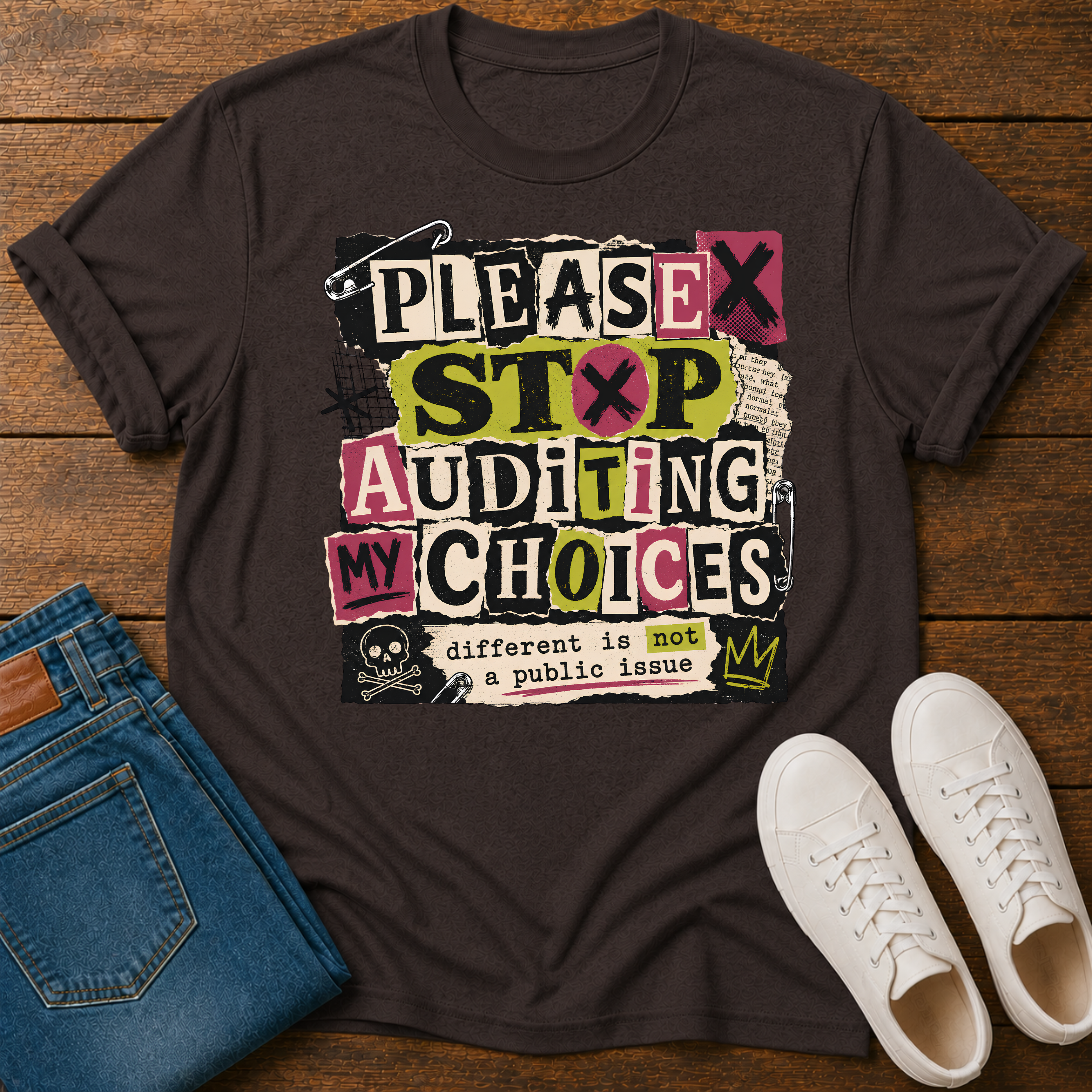 A dark brown graphic t-shirt laid flat on a wooden surface, styled with folded blue jeans and white sneakers. The t-shirt features a colorful, distressed graphic that reads 'PLEASE STOP AUDITING MY CHOICES' with a small skull and the text 'different is not a public issue'.