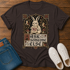 A dark brown graphic t-shirt laid flat on a wooden surface, styled with blue jeans and white sneakers. The t-shirt features a vintage-style illustration of a sad rabbit holding a sign that says 'NO' surrounded by birds and mushrooms, with the text 'ASK someone ELSE'.